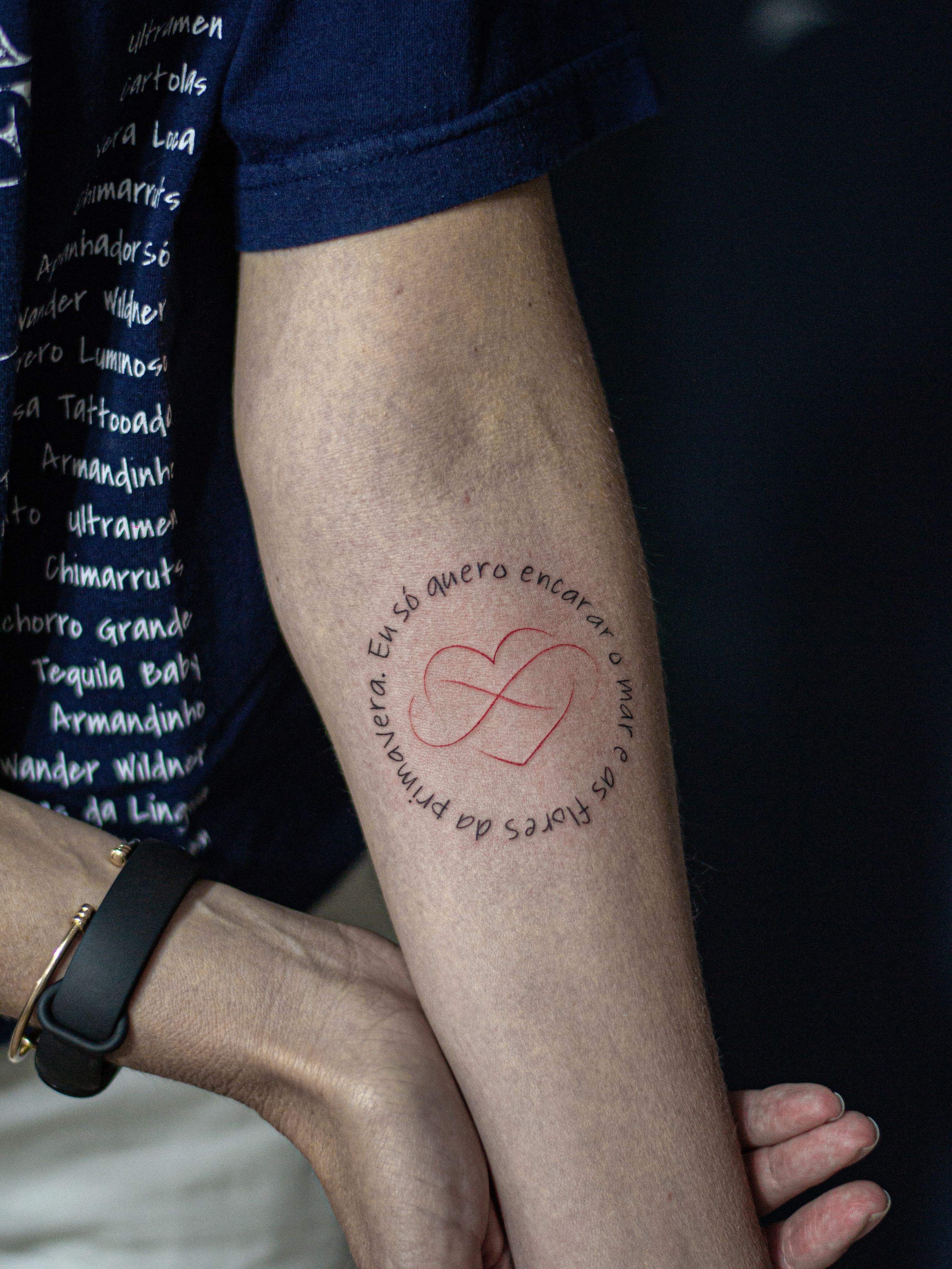 Minimalist tattoo made in fineline on the forearm area, representing a broken heart design, intertwined with the infinity symbol and with the phrase "I just want to face the sea and the spring flowers".