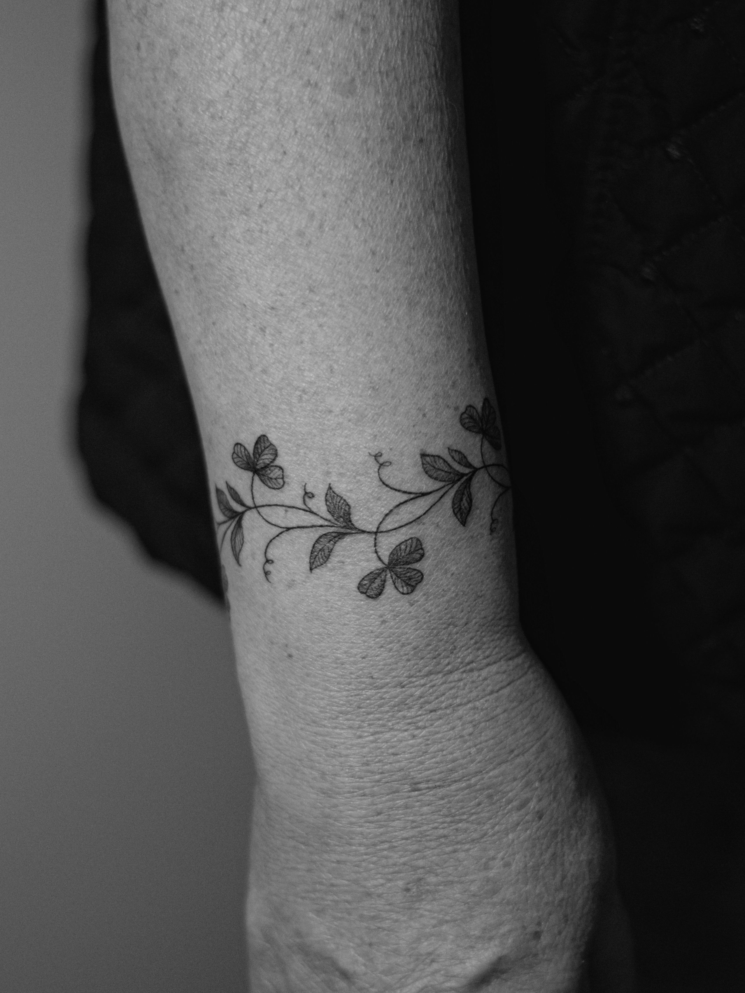 Minimalist tattoo made in fineline on the forearm area, in bracelet format, representing a branch of clover leaves.