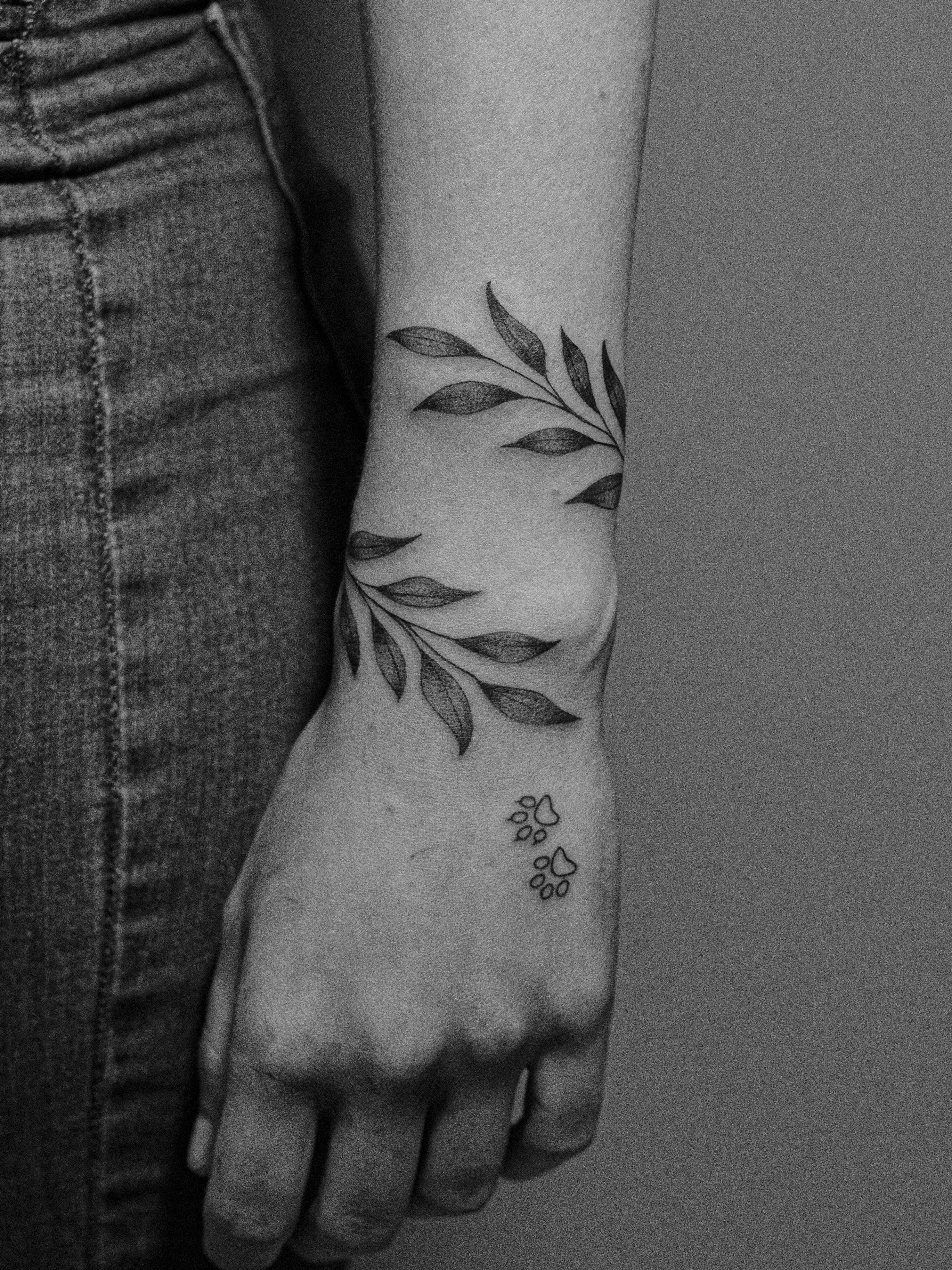 Minimalist tattoo made in fineline on the forearm/wrist area, representing a branch of leaves. Further down, on the hand, two paw prints, one cat and one dog.