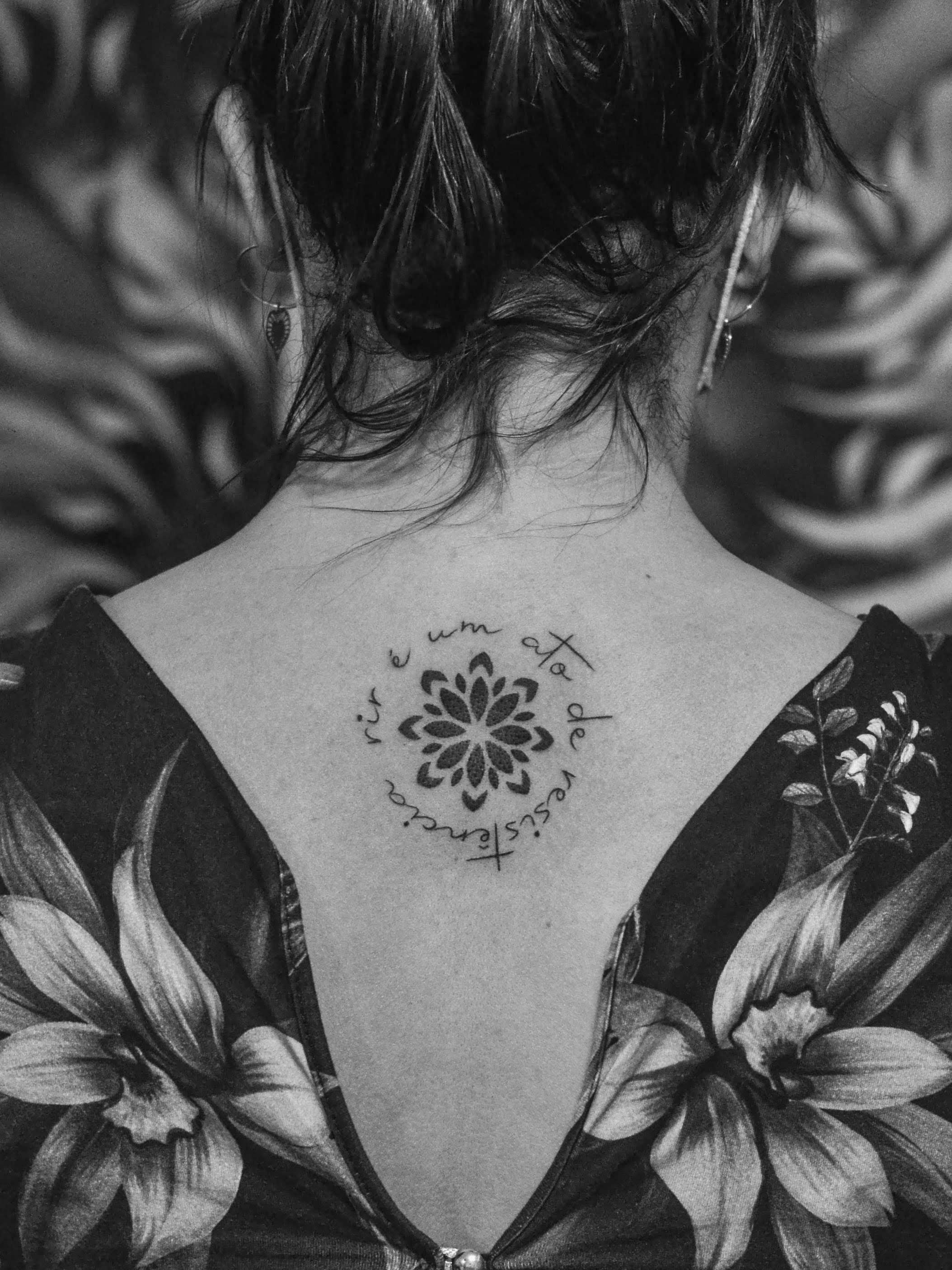 Minimalist tattoo made in fineline and dotwork on the back, on the spine area, representing a mandala flower and the phrase "laughter is an act of resistance", around it; in a circular shape.