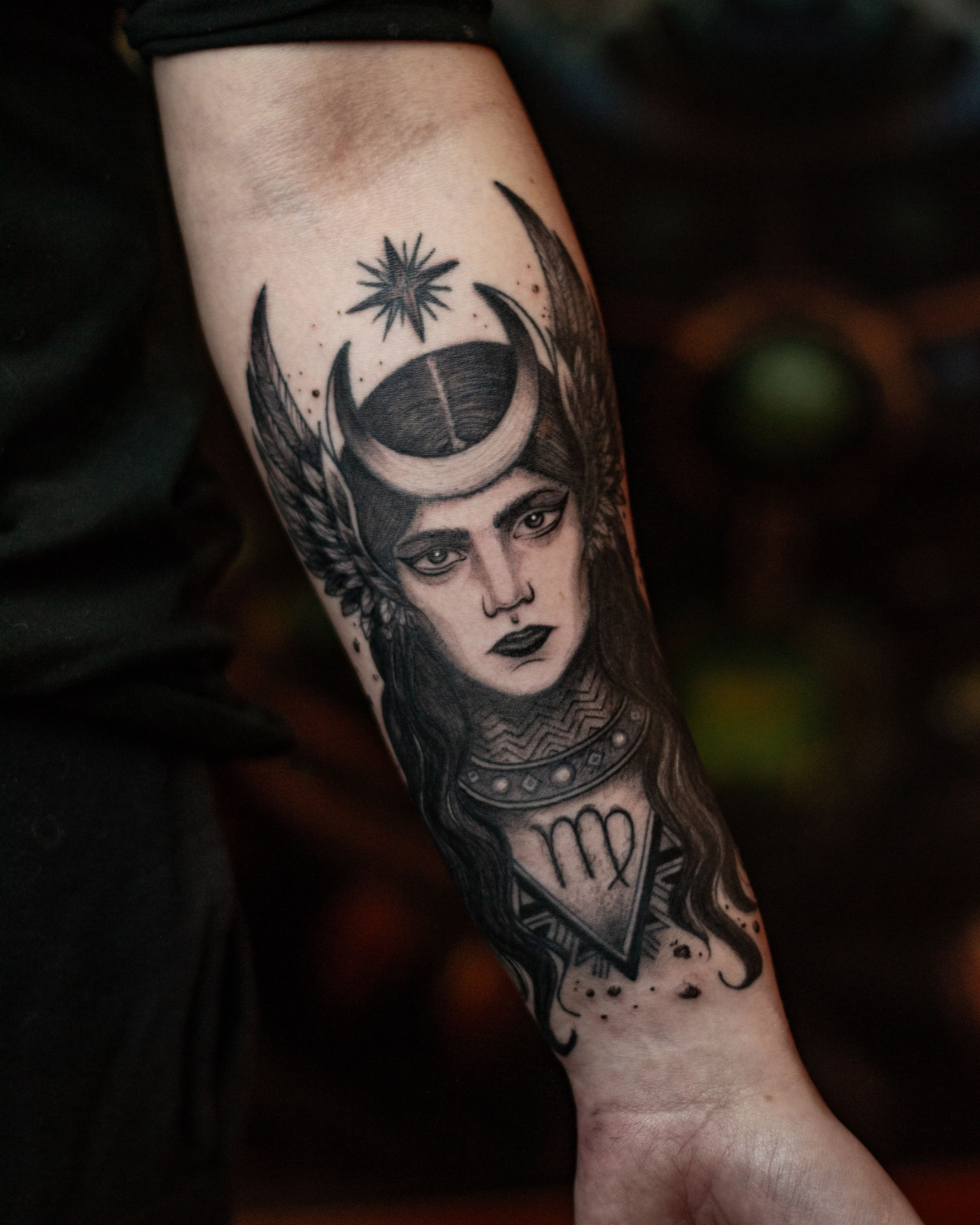 Detailed tattoo of a mystical female figure, adorned with a lunar crescent and side wings. Above, a guiding star; below, the Virgo symbol. The deep gaze and striking features evoke spirituality, power, and cosmic connection.